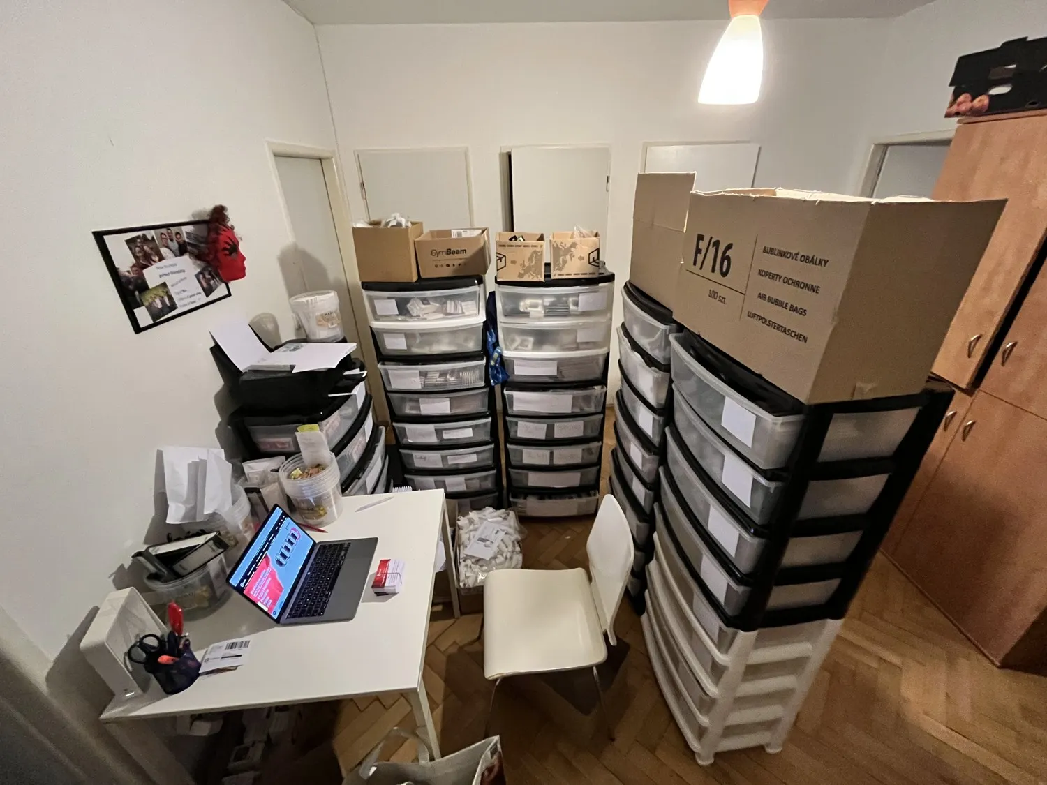 Izba v byte premenená na sklad e-shopu: stohy plastových šuplíkov, krabice s obálkami a baliaci stôl